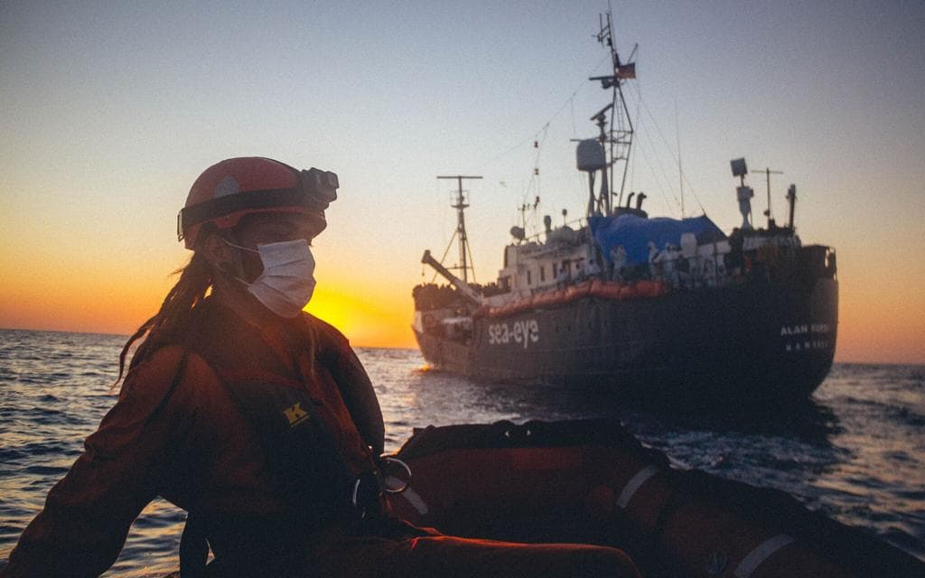 Sun sets behind Sea-eye boat, in the foreground a masked person sits aboard a small boat